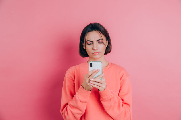 表情年轻女子在阅读手机短信时感到悲伤 不开心 愤怒 背景是粉色的室内信息成人