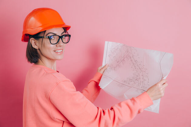年轻在粉色背景下微笑的女工建筑工人建筑头盔人工作保护
