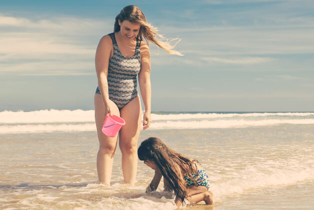 海滩快乐的妈妈和小女儿站在足踝深的海水和湿沙里 把贝壳捡进桶里水桶站发现