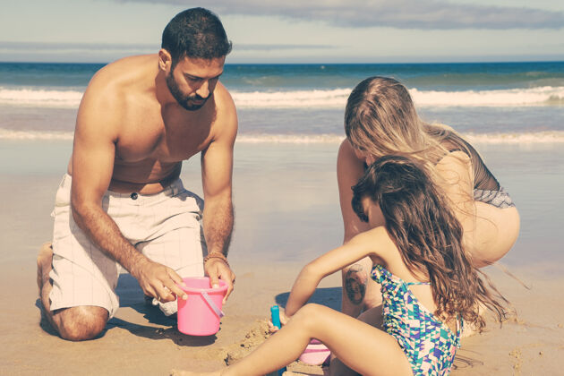 水年轻的父母和可爱的小女孩在沙滩上嬉戏玩耍 用玩具铲 桶和碗挖土肖像享受玩