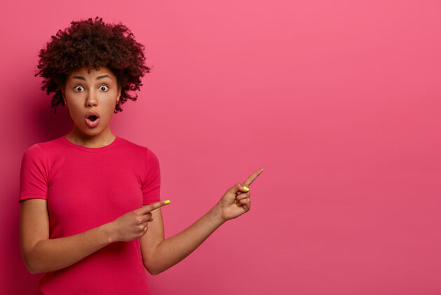 无语惊讶的非裔美国女人指了指 为你的广告显示空白 震惊于难以置信的报价 张开嘴 穿着随意 隔离在粉红色的墙壁天哪 看看这个年轻种族惊艳