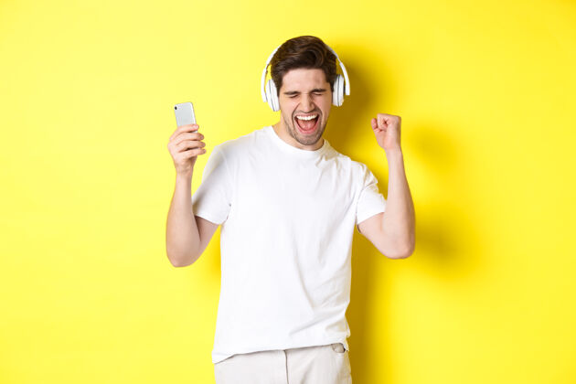 耳机快乐的男人戴着耳机跳舞 听音乐 拿着手机 站在黄色的背景下黄色模特帅哥