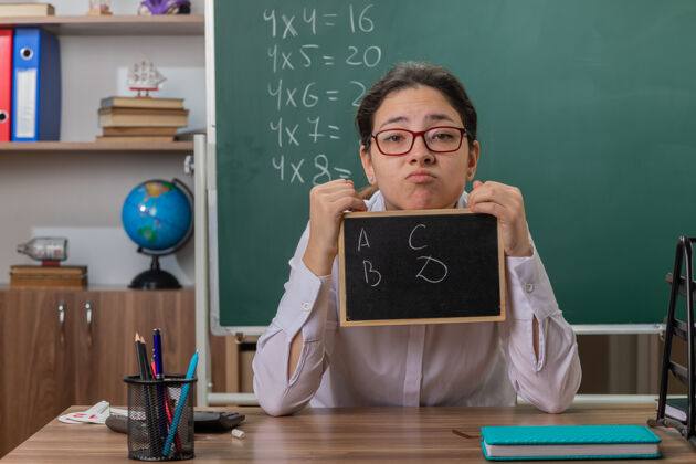 教室年轻的女老师戴着眼镜 拿着小黑板 用困惑的表情看着前面 坐在教室黑板前的课桌上讲解课文课桌黑板女