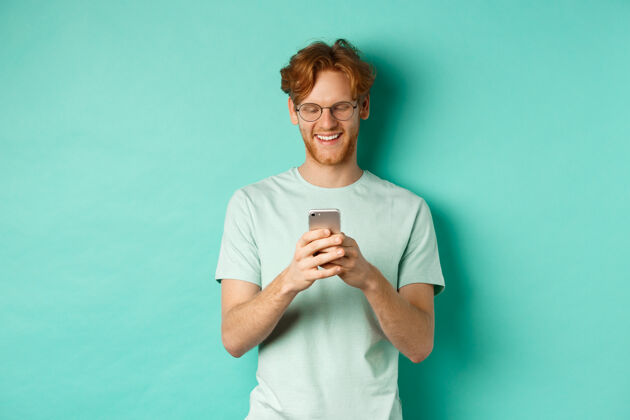 男戴着眼镜 头发凌乱的帅哥在看手机短信 微笑着看着屏幕 站在薄荷色的背景上沟通胡子人