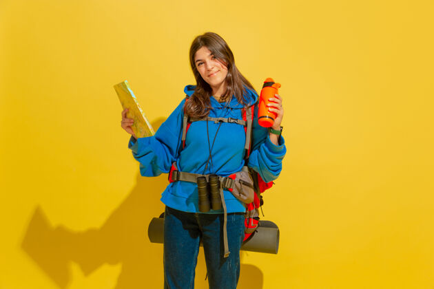 欢呼黄色摄影棚墙上 一个带着包和望远镜的快乐的年轻旅游女孩的画像人快乐女人