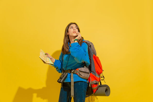 年轻人黄色摄影棚墙上 一个带着包和望远镜的快乐的年轻旅游女孩的画像人光明欢呼