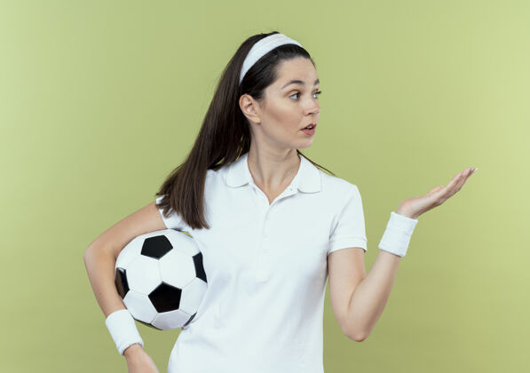 手臂戴着头巾拿着足球的年轻健身女士站在轻质的墙壁上 她的手臂看起来很惊讶年轻光头带
