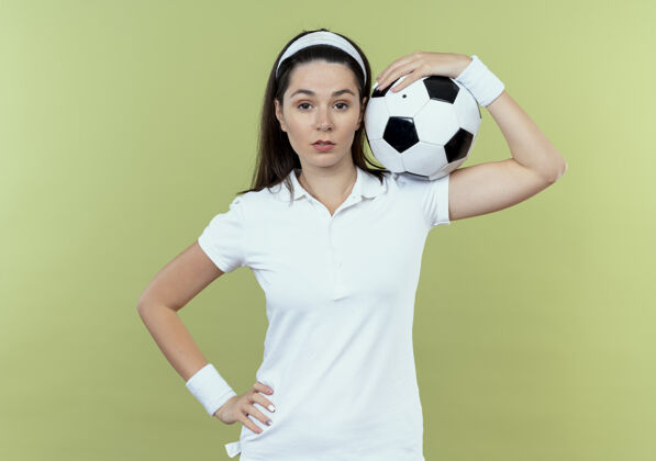 年轻戴着头巾的年轻健身女士肩上扛着足球 自信地站在轻盈的墙上举行表达轻
