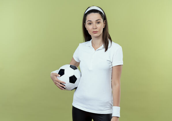 球戴着头巾的年轻健身女士手持足球 表情严肃自信地站在轻盈的墙上女子严肃表情