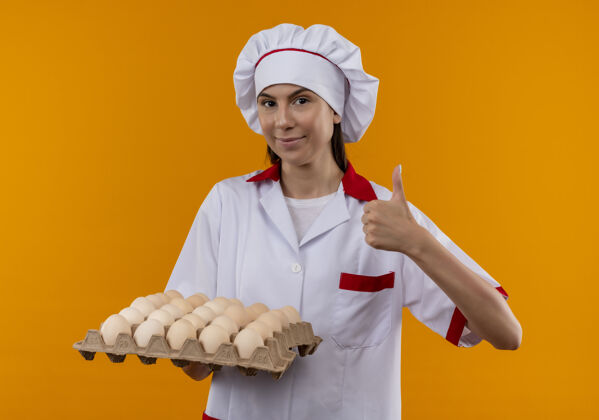 向上穿着厨师制服的年轻快乐的白人厨师女孩拿着一批鸡蛋 竖起大拇指 孤立地站在橙色的空间里复制空间橙色复制女孩