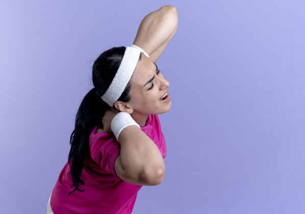穿戴着头带和腕带的年轻的白人运动女性站在一边 把脖子放在后面 在紫色的空间和复制空间隔离疼痛立场侧