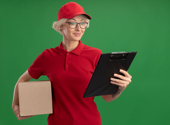 立场年轻的送货员身穿红色制服 戴着帽子 戴着眼镜 手里拿着纸板箱和剪贴板 面带微笑地看着它站在绿色的墙上穿眼镜举行