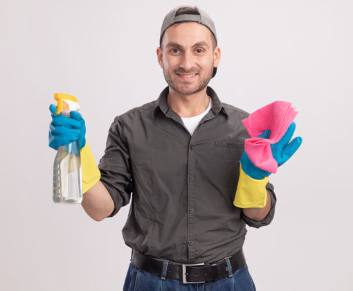 举行年轻的清洁工穿着休闲服 戴着橡胶手套 戴着帽子 手里拿着喷雾瓶和抹布 站在白色的墙上 脸上洋溢着幸福和积极的微笑男人抹布立场