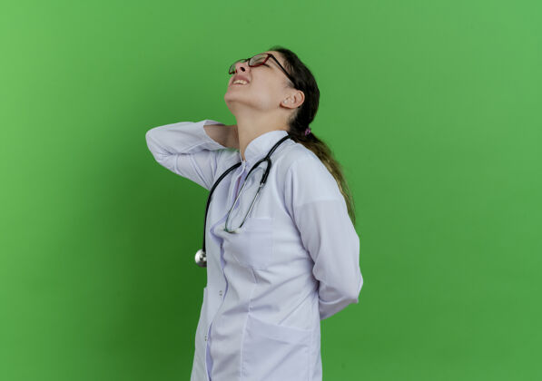听诊器疼痛的年轻女医生穿着医用长袍 戴着听诊器和眼镜站在侧视图中 将手放在脖子后面和后面 隔离在绿色墙壁上 留有复印空间视图年轻长袍