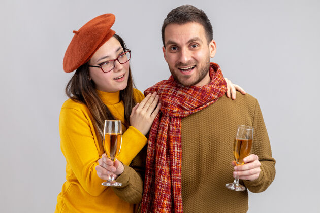 站立年轻漂亮的夫妇戴着贝雷帽的快乐的男人和微笑的女人 戴着香槟酒杯 快乐地恋爱 站在白墙上庆祝情人节情人节日子一起