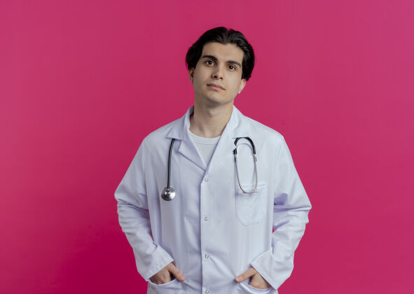 长袍年轻的男医生穿着医用长袍和听诊器把手放在口袋里 隔离在粉红色的墙上 留有复印空间听诊器医生手