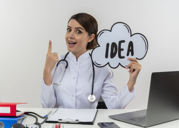 穿令人印象深刻的年轻女医生穿着医用长袍和听诊器坐在办公桌旁 手里拿着医疗工具和笔记本电脑 手里拿着创意泡泡 举起手指孤立在白墙上工具创意女性