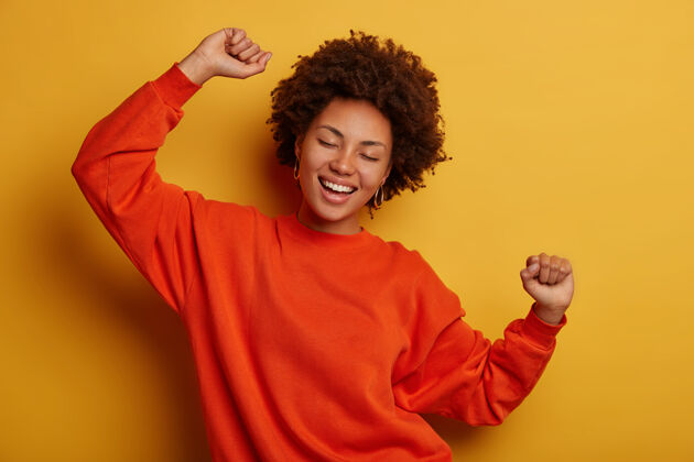 非洲快乐的黑发女人有乐趣 举手跳舞 穿着休闲套头衫 在黄色背景下欢呼 得到晋升或批准 庆祝胜利女士女性室内