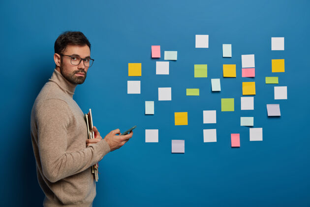 日记自信的男职工在蓝色的墙上整理五颜六色的笔记 写项目创意 用手机 上网搜索信息安排记事本贴纸