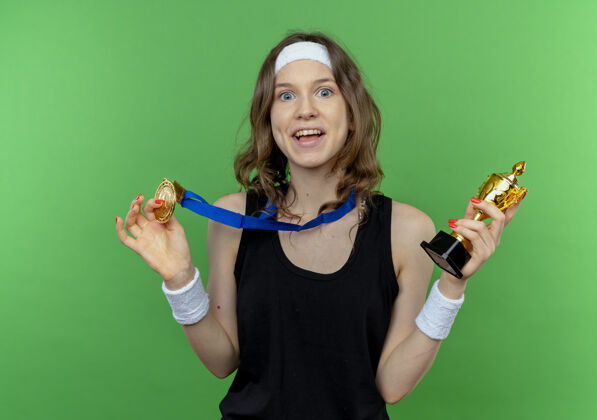 运动装身穿黑色运动服的年轻健身女孩戴着头带 脖子上挂着金牌 手里拿着奖杯 为绿色感到高兴和兴奋快乐金牌健身