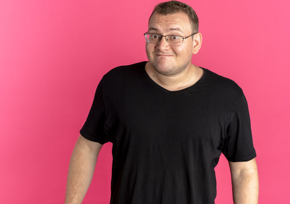 粉色戴眼镜的超重男子穿着黑色t恤困惑地微笑着 而不是粉红色站男人T恤
