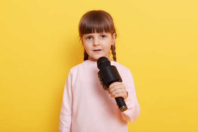 卡拉OK可爱的小女孩 带着黄色背景的麦克风 一边用麦克风说话 一边看着摄像机 食指指向一边为广告或促销文本复制步调麦克风声音肖像