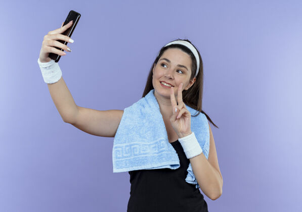 健身戴着头巾脖子上围着毛巾的年轻健身女士看着智能手机屏幕自拍展示胜利歌唱微笑着站在蓝色的墙上毛巾显示女人