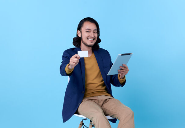 肖像亚洲人坐在明亮的蓝色椅子上 愉快地微笑着出示信用卡和使用数字平板电脑折扣媒体Pad