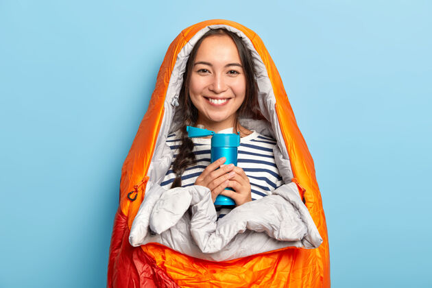 请快乐的女人留着长长的辫子 站在睡袋里 拿着热饮料的保温瓶 喜欢旅游和徒步旅行饮料女性黑发