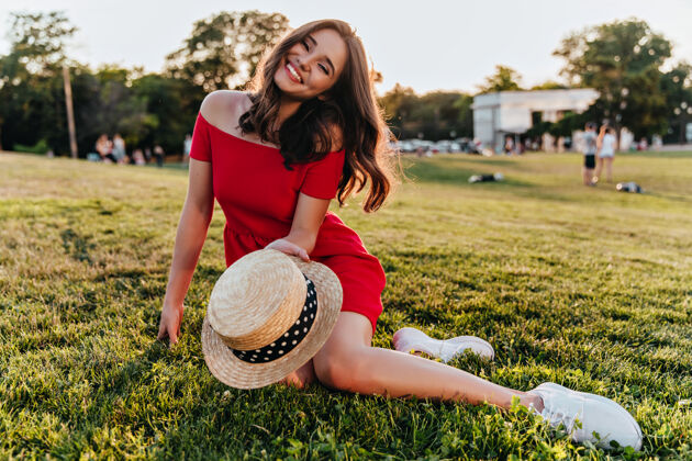 美丽正派的黑发女士坐在草地上 手里拿着夏帽优雅的白人女孩 在公园里和划船的人在一起时尚优雅休闲