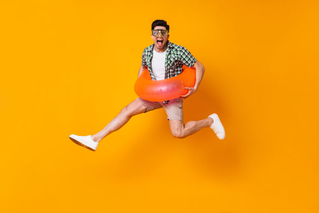 文件年轻淘气的男人穿着夏天的行头 戴着潜水面具 在橙色的空间里用充气圈跳跃鸡尾酒包衬衫