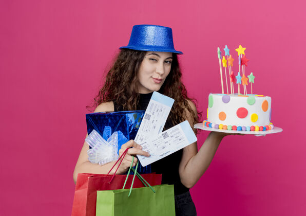 微笑一位年轻漂亮的女士 卷发 戴着节日礼帽 手里拿着生日蛋糕礼盒和机票 微笑着 眨眼着站在粉色的墙上的生日派对概念抱着蛋糕卷发