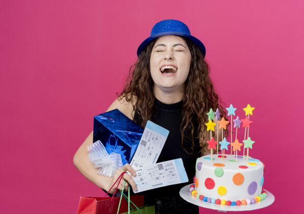 年轻一位年轻漂亮的女士 卷发 戴着节日礼帽 手里拿着生日蛋糕礼盒和机票 站在粉色的墙上 快乐而兴奋的生日派对概念帽子抱着蛋糕