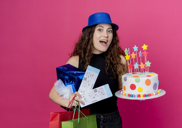 卷发一位年轻漂亮的女士 卷发 戴着节日礼帽 手里拿着生日蛋糕礼盒和机票 站在粉色的墙上 快乐而兴奋的生日派对概念抱着飞机女人
