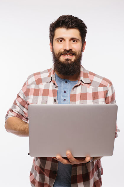 快乐专注的年轻留着胡子的男子戴着眼镜 穿着衬衫 用笔记本电脑隔着白墙互联网成人笔记本电脑