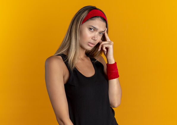 运动装穿着黑色运动服 戴着红色头巾的年轻健身女孩用手指着太阳穴 站在橘色的墙上使劲儿头带立场健身
