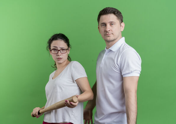 运动一对年轻的运动型情侣 拿着棒球棒站在她男朋友旁边 严肃的脸站在绿色的墙上棒球男朋友蝙蝠