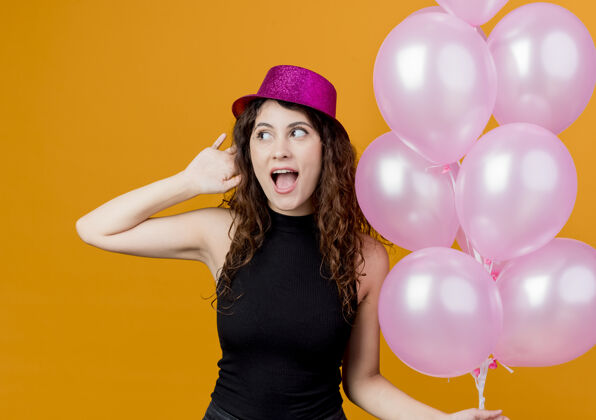 卷发一个年轻漂亮的女人 卷发 戴着节日礼帽 手里拿着一堆气球 快乐而兴奋地微笑着 站在橘色的墙上 欢欣鼓舞地庆祝生日欢呼气球举行