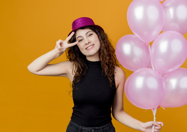 气球一位年轻漂亮的女士 卷发 戴着节日礼帽 手里拿着一束气球 开心而积极地微笑着 站在橙色的墙上 欢快地展示着v字生日派对的概念Vsign帽子积极