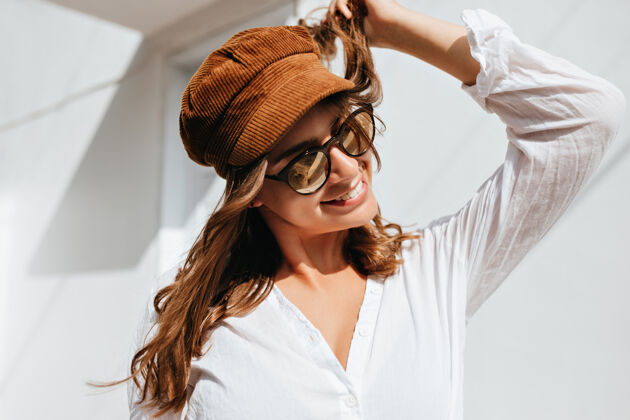 短发年轻女子把玩她的头发和微笑女孩穿着白色衬衫 棕色帽子和太阳镜对白色的建筑物摆姿势城市温暖裤子