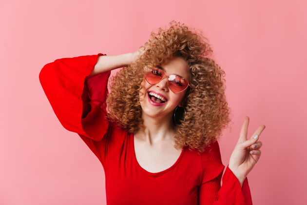 头发开朗的金发卷发女子笑着在粉色空间展示和平标志戴着太阳镜和红色上衣的女士的快照享受红色衬衫表情