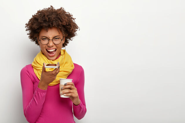 室内工作室拍摄的愤怒恼怒的非洲发型女人 通过智能手机打电话 喝外卖咖啡 戴眼镜 粉色高领毛衣和黄色围巾 隔着白墙围巾罗西脸