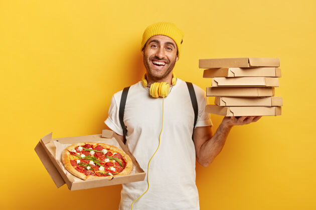 工作送货概念男人披萨经销商拿着一堆纸板箱 在打开的集装箱里展示美味的快餐 当快递员 戴着黄色帽子和白色t恤 用耳机听音频携带送货背包