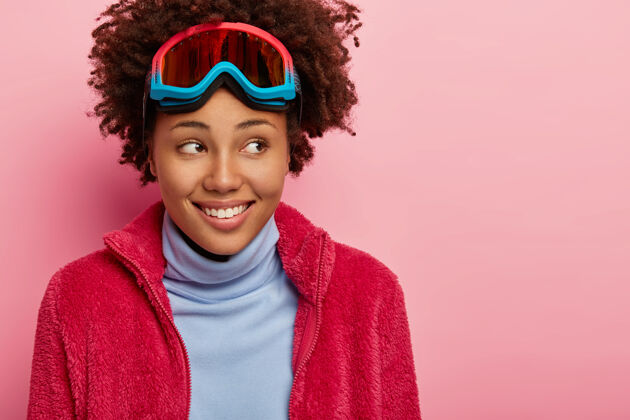 欢呼高兴的卷发女性穿着冬装 前额戴着滑雪镜 高兴地看着一旁 模特们翻过粉色的墙壁滑雪板眼镜装备
