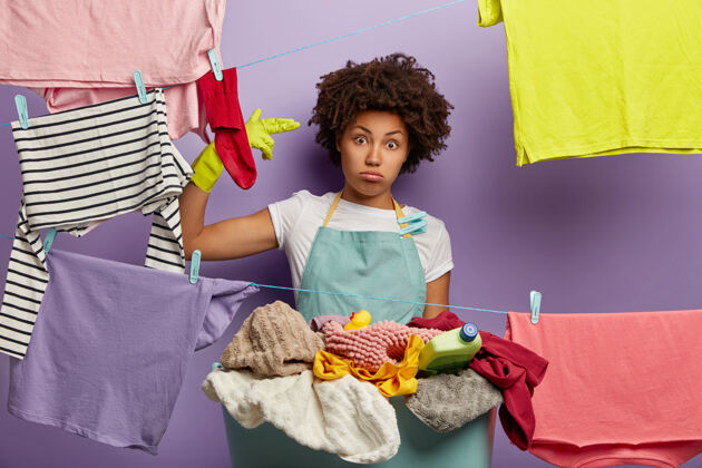 不安沮丧悲伤的女人做出的姿态 在家里有很多工作 穿着休闲围裙 周末洗衣服 挂干净的衣服 在室内摆姿势衣夹不满不快乐
