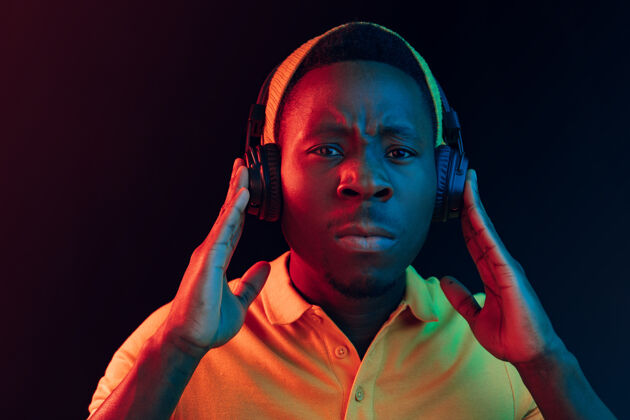 享受年轻帅气严肃悲伤的潮人戴着耳机在黑色霓虹灯下听音乐非洲男性严肃
