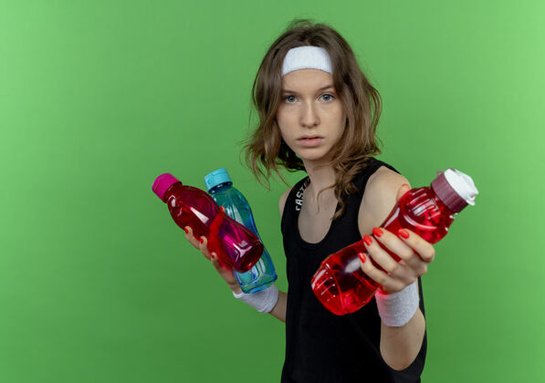 瓶子穿着黑色运动服的年轻健身女孩 头上戴着一条带子 手里拿着几瓶水 在绿色的墙上放着一瓶水年轻运动装女孩