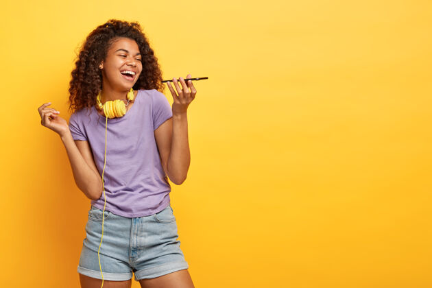 室内摄影棚拍摄的快乐的年轻女性与非洲发型摆在黄色的墙壁通话欣喜若狂电话