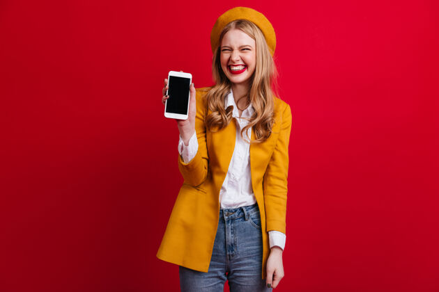 女人幸福的法国女人拿着智能手机和空白屏幕前视图优雅的金发女孩在贝雷帽隔离在红墙年轻人空白屏幕数码设备
