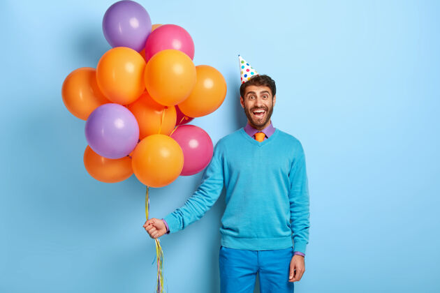 姿势带着生日帽和气球的微笑的家伙穿着蓝色毛衣摆姿势成人男性帽子